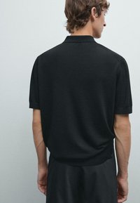 Černé polo tričko s krátkým rukávem vyrobené z hladké tkaniny, s klasickým límečkem a úhledně složenými rukávy, viděné z pohledu zezadu.
