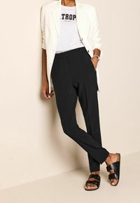 Pantalon noir ajusté avec une texture lisse, associé à un t-shirt graphique gris clair, un blazer blanc et des sandales noires.