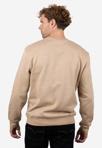 Sudadera de algodón beige con cuello redondo, puños y dobladillo acanalados, y un sutil patrón tonal en la parte trasera. Combinada con pantalones vaqueros oscuros.