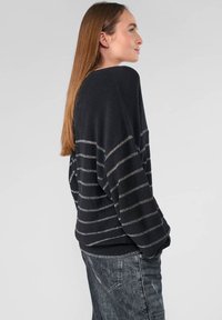 Zwarte gebreide trui met brede mouwen, voorzien van horizontale zilveren strepen, geribbeld zoom en een losse pasvorm. Gecombineerd met grijze jeans.