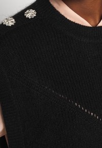 Un pull en tricot noir avec deux embellissements en argent décoratifs sur les épaules et une chemise à col rose clair en dessous.