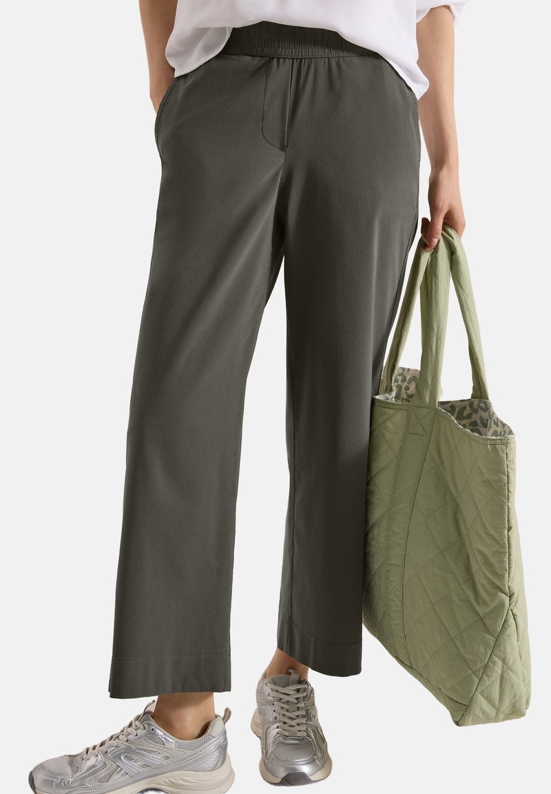 Personne portant un pantalon ample vert olive et des baskets argentées, tenant un sac cabas vert matelassé avec une doublure à motifs.
