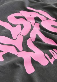 Sort tekstil med en rynket overflade, der har store, uregelmæssige pink bogstaver. Designet fremstår grafisk og dristigt med en mat finish.