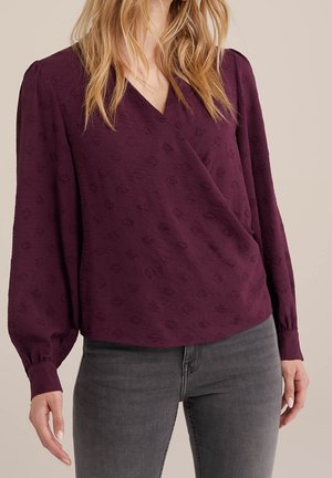 Vrouw met een langermige, gestructureerde pruimenblouse met overslag aan de voorkant, gecombineerd met donkergrijze spijkerbroek.