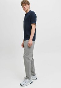 Giovane uomo in piedi di lato, indossando una polo blu scuro a maniche corte, pantaloni grigi chiari e scarpe da ginnastica bianche su uno sfondo neutro.