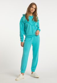 Set di felpa e pantaloni jogger color turchese in tessuto morbido, con pompon decorativi sulla felpa. I pantaloni hanno cavigliere a coste e una vestibilità rilassata.