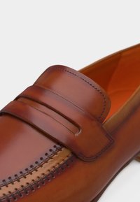 Καφέ δερμάτινο μοκασίνι με διακοσμητικό ράφτη και λείο λουράκι στο πάνω μέρος, που δείχνει λεπτομερή υφή και τεχνική.