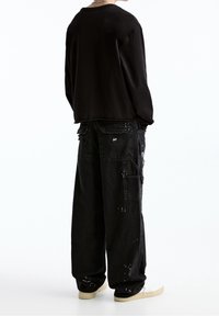 Camisa negra de manga larga, pantalones cargo negros de corte holgado con múltiples bolsillos y detalles de salpicaduras de pintura, combinados con zapatos de color claro.