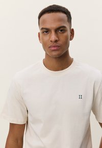 T-shirt en coton couleur crème avec col rond et manches courtes. Présente un petit "II" vert brodé sur la poitrine. Texture lisse.