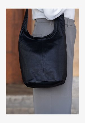 Bolso de hombro de piel negra con una textura suave y lisa, con parte superior curva y base redondeada, presentado junto a unos pantalones de color claro.
