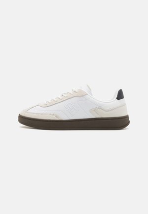 Zapatilla blanca con parte superior de ante y cuero, un acento crema, una suela de goma marrón y un logo en el lado con un talón acolchado.