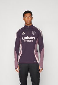 Langärmliges Sportoberteil in lila mit rosa Akzenten, hohem Kragen, Halbreißverschluss vorne und weißem Sponsorlogo. Aus leichtem Material gefertigt.