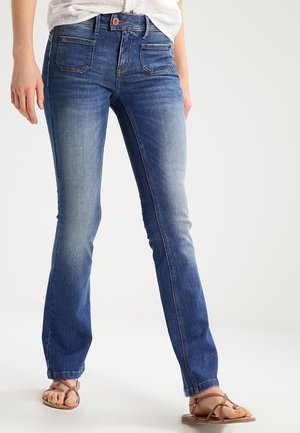 Person iført blå straight-leg jeans, hvid top og brune sandaler med remme står mod en ensfarvet lys baggrund.