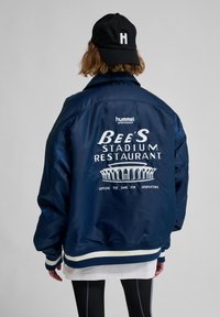 Navy satin bomber jacket with ribbed cuffs and hem featuring white embroidered text and a stadium graphic on the back, designed by Hummel.
