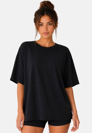 Jeune femme aux cheveux bruns attachés en chignon, portant un t-shirt noir oversize et un short noir sur un fond clair uni.