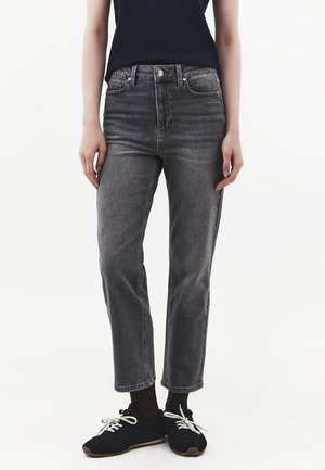 Person iført højttaljede falmede sorte jeans, sorte sokker, sorte sneakers med hvide snørebånd og en mørk ærmeløs top mod en hvid baggrund.