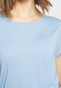 Jasnoniebieska koszulka z krótkim rękawem o okrągłym dekolcie i miękkiej, gładkiej teksturze. Posiada luźny krój z delikatnymi detalami po bokach.