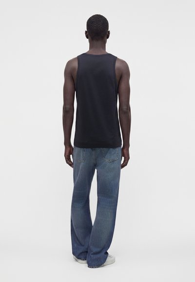 Άνδρας με μαύρο αμάνικο πουκάμισο, μπλε φαρδιά τζιν και άσπρα παπούτσια, που στέκεται γυρισμένος πλάτη σε ένα απλό φωτεινό φόντο.