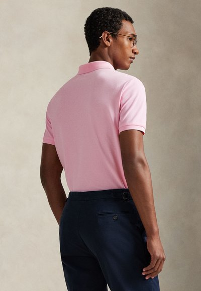 Πόλο μπλούζα σε ανοιχτό ροζ βαμβάκι, με κλασικό γιακά και κοντά μανίκια, συνδυασμένη με σκουρόχρωμο ναυτικό μπλε παντελόνι. Καθαρός και μοντέρνος σχεδιασμός.