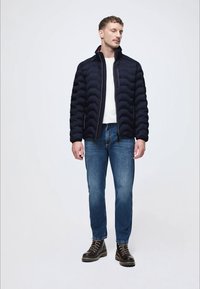 Επενδεδυμένο ναυτικό μπουφάν με ψηλό γιακά, σε συνδυασμό με λευκό πουκάμισο και μπλε τζιν. Φορά σκούρα δερμάτινα μποτάκια με χοντρή σόλα.