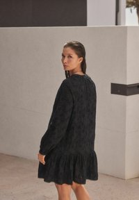 Robe noire brodée avec des manches longues, un col rond et un ourlet à volants. Présente des motifs floraux et de petites découpes, tissu texturé.