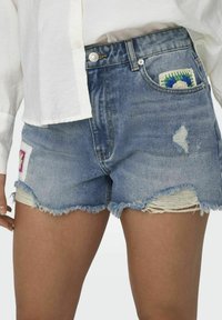 Shorts de mezclilla con dobladillo deshilachado, lavado azul claro y parches coloridos en los bolsillos delanteros. Detalles desgastados presentes.