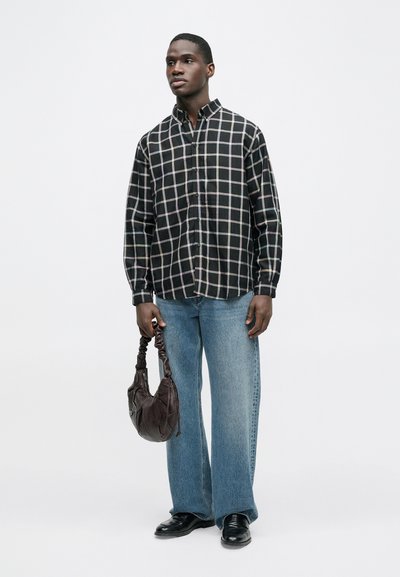 Πουκάμισο με ασπρόμαυρο καρό, συνδυασμένο με ανοιχτό μπλε παντελόνι με φαρδιά πόδια, κρατώντας μια καφέ σκούρα τσάντα με υφή. Μαύρα παπούτσια.