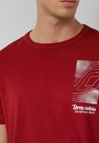 Mężczyzna w czerwonym t-shircie z okrągłym dekoltem, na którym widoczne są białe linie topograficzne oraz napis "Terra visible NA NATURAL PATH" na piersi.