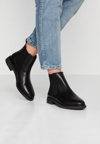 Bottines noires en cuir avec des panneaux latéraux élastiques et une semelle en caoutchouc texturée, assorties à un jean léger bleu clair retroussé.