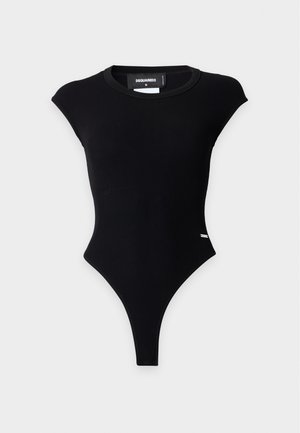Femme aux cheveux noirs portant une veste grise, un body noir avec des découpes latérales, un jean bleu foncé et des talons ouverts noirs, debout les mains dans les poches.