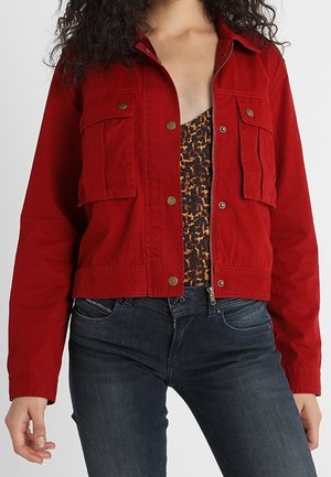 Vrouw draagt een rode cropped jas met knoopzakken over een luipaardprint top en donkerblauwe spijkerbroek.