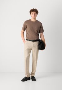 Brown short-sleeve T-shirt, beige linen trousers, black belt, and black shoes; holding a black jacket. Smooth fabric textures, relaxed fit.