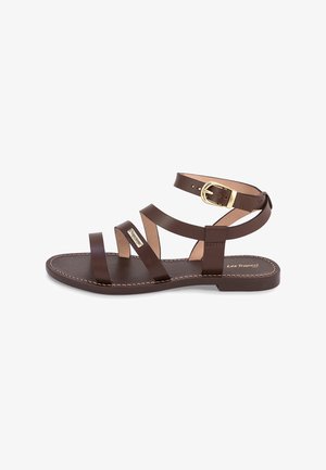 Sandales plates en cuir marron avec trois larges brides et une fine sangle à la cheville, dotées d'une boucle dorée et d'une semelle claire contrastante.