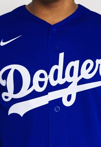 Modrá baseballová dres vyrobená z texturované látky, s velkým bílým nápisem "Dodgers" a dvěma modrými knoflíky na límci. Logo Nike na rameni.