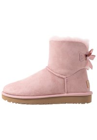 Bottine en daim rose clair avec une doublure en fourrure douce, un bout arrondi, et un détail de nœud décoratif à l'arrière, complétée par une semelle en caoutchouc beige.