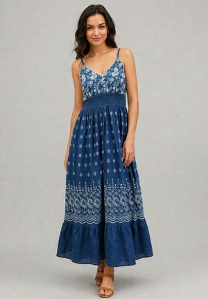 Femme portant une robe maxi bleue sans manches avec des motifs floraux blancs et paisley, debout avec une main détendue le long du corps.