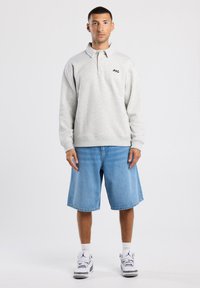 Polo a maniche lunghe grigio chiaro con colletto a bottoni e piccolo logo nero, abbinato a pantaloncini di jeans azzurri e scarpe da ginnastica bianche.