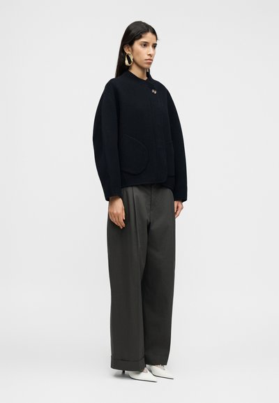 Μαύρο μάλλινο σακάκι με στρογγυλή λαιμόκοψη, κούμπωμα μπροστά με κουμπιά και δύο μεγάλες τσέπες, σε συνδυασμό με άνετο γκρι παντελόνι και λευκά μυτερά παπούτσια.