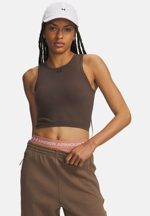 Vrouw met een witte pet, bruin mouwloos crop top, bruine joggingbroek en een zichtbare roze Under Armour tailleband, staand tegen een effen achtergrond.