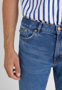 Keskitummansiniset farkut, joissa klassinen leikkaus, viiden taskun malli, oranssiompeleet ja kuparinväriset yksityiskohdat.