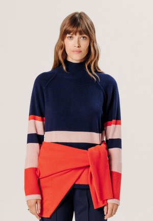 Femme aux cheveux mi-longs portant un pull bleu marine avec des rayures roses et rouges, et un pull rouge vif noué autour de la taille.