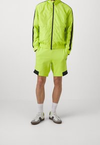 Neon grøn tracksuit med sorte detaljer, der har en lynlåsjakke og matchende shorts. Materialet er glat og atletisk. Hvide sokker og sportssko.