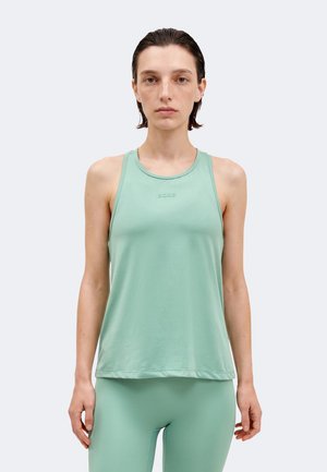 Νεαρή γυναίκα με κοντά βρεγμένα μαλλιά φοράει ανοιχτοπράσινο αμάνικο τοπ και ταιριαστά κολάν, κοιτάζοντας μπροστά σε μονόχρωμο φόντο.