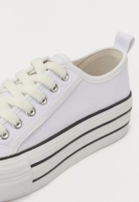 Sneaker bianca in tela con suola platform spessa, caratterizzata da righe nere e bianche, punta tonda e lacci bianchi piatti attraverso occhietti in metallo.