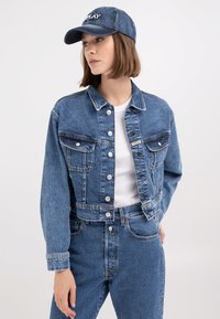 Kék farmerdzseki, cropped fazonnal, hosszú ujjakkal, gombos záródással, mellzsebekkel és kontrasztos varrással, egy azonos színű sapkával párosítva.