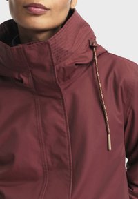 Veste bourgogne avec un col haut, présentant une surface texturée et un cordon de serrage avec une pointe beige. Détails de couture visibles le long du col.