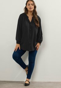 Camicia nera a maniche lunghe con bottoni, dal taglio comodo, con scollatura a V e polsini arricciati, abbinata a jeans skinny blu e ballerine nere.
