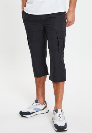 Homme portant un pantalon cargo capri noir avec des poches latérales et des baskets blanches, bleues et jaunes sur fond blanc.