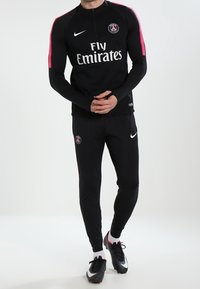 Schwarzer sportlicher Trainingsanzug mit einem langärmligen, mit Reißverschluss versehenen Oberteil, das pinkfarbene Akzentärmel, das Nike-Logo und den Text "Fly Emirates" aufweist. Enthält eine enganliegende Hose.