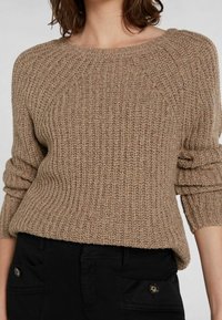 Maglione marrone lavorato a maglia con una texture a costine, scollatura ampia e maniche arrotolate. Abbinato a pantaloni neri con dettagli a bottoni.